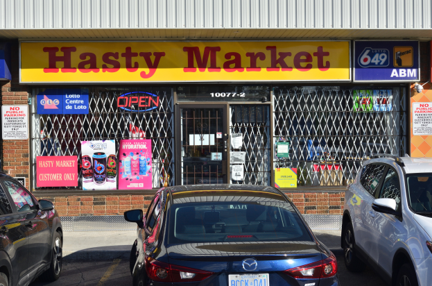 Ein belebter Hasty Market in Toronto mit Autos auf der Straße davor, mit Glaswänden und Türen mit Text enthaltenden Postern.