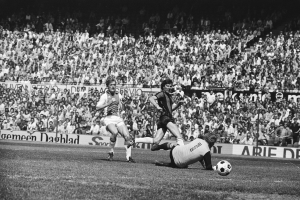 Schwarzes Foto von Männern, die auf einem Feld Fußball spielen, mit Zuschauern im Hintergrund und einem Banner 'Bundesliga 1978-79 Ajax 1-1 Hannover 96'.
