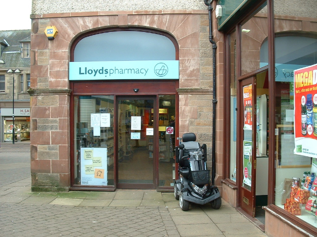 Eine Apotheke mit einem vor ihr geparkten Fahrzeug und einem Gebäude links daneben.