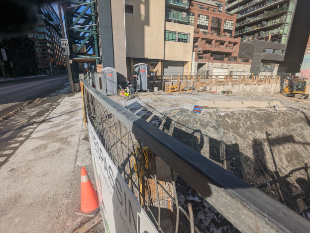 Eine Baustelle mit einem Zaun und einer Verkehrskegel im Vordergrund, eine Straße links, Gebäude im Hintergrund, ein Baumaschinenfahrzeug und ein Müllcontainer.