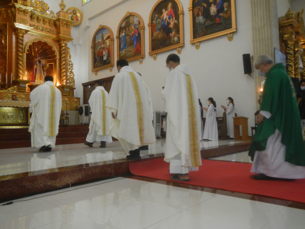 Eine Gruppe von Priestern geht eine rote Teppich in einer Kirche entlang, mit Menschen, die auf dem Boden vor ihnen und auf der rechten Seite stehen, und Fotorahmen an der Wand im Hintergrund.