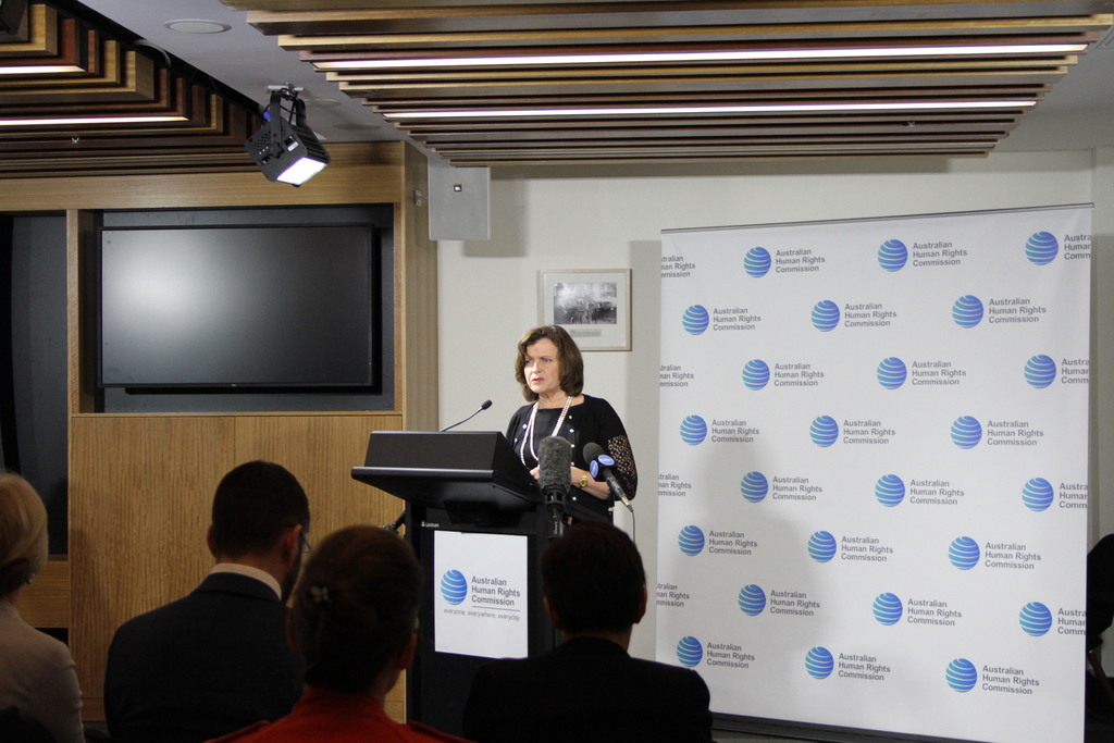 Eine Frau steht auf einer Bühne und spricht in ein Mikrofon, vor ihr sitzen Menschen auf Stühlen, an der Wand hängt ein Banner, ein Fernseher steht an der Holzwand und an der Decke leuchten Lampen.