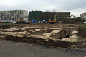 Große Erdaufschüttung auf einer Baustelle umgeben von Baggern, Fahrzeugen, Gras, Bäumen und Gebäuden mit Fenstern in einem bewölkten Himmel, mit einer Straße unten.