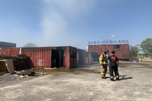 Zwei Feuerwehrleute in Helmen vor einem raucherfüllten Gebäude, einer hält ein Rohr, mit verstreuten Trägern und einem Namensschild im Hintergrund; Trees und ein klarer Himmel sind im Hintergrund zu sehen.