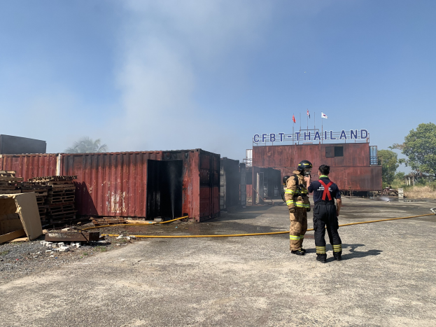 Zwei Feuerwehrleute in Helmen vor einem raucherfüllten Gebäude, einer hält ein Rohr, mit verstreuten Trägern und einem Namensschild im Hintergrund; Trees und ein klarer Himmel sind im Hintergrund zu sehen.