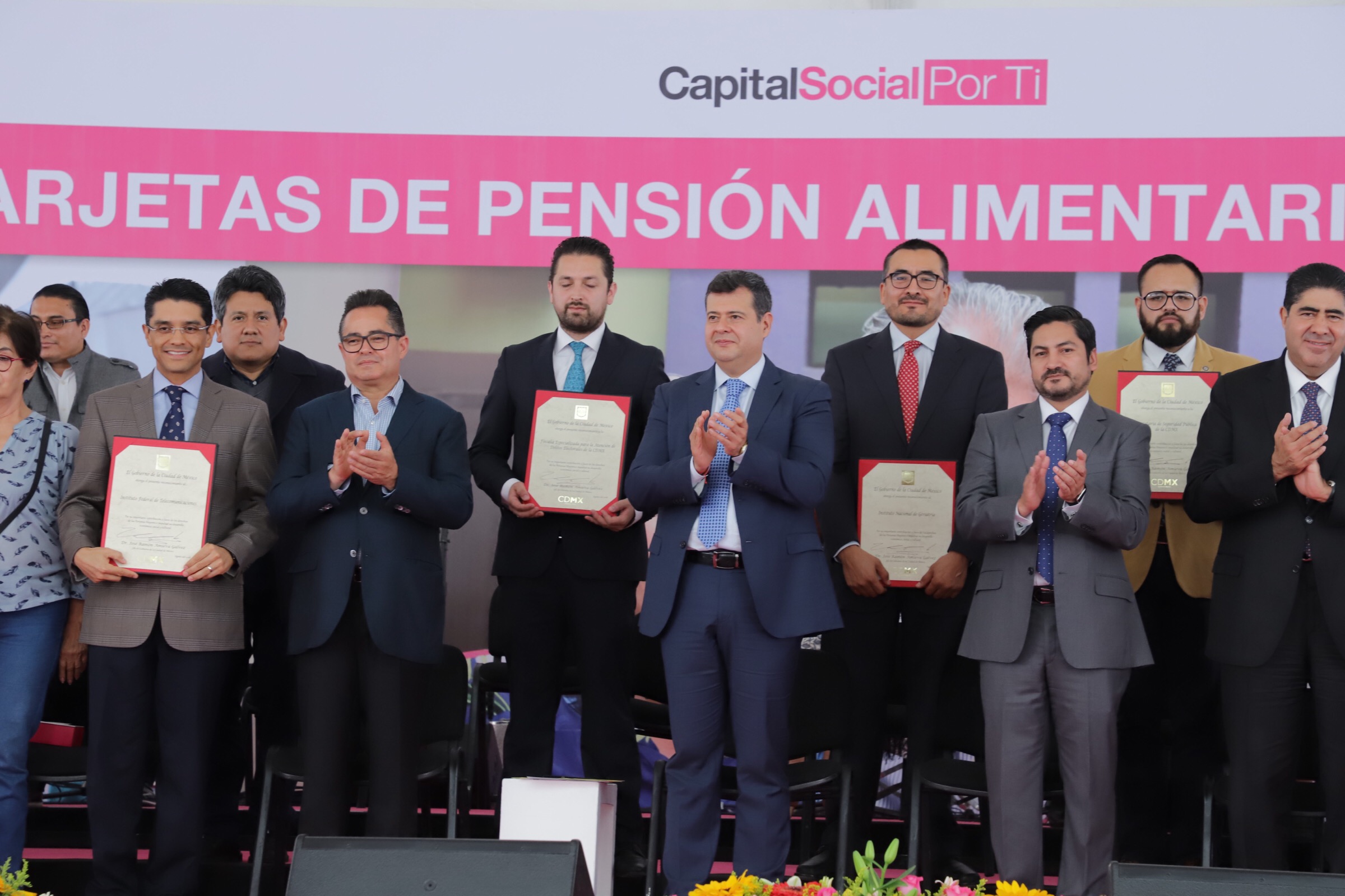 Eine Gruppe von Menschen auf einer Bühne, die klatschen und Zertifikate halten, mit Stühlen in einem Halbkreis darum und einem Banner mit der Aufschrift "Capital Social for Ti" im Hintergrund, sowie Blumensträuße und Redner am Boden.