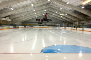 Große Indoor-Eisfläche mit Deckenbeleuchtung, Werbetafeln, einer Wand, Treppen mit Geländern und einigen Menschen, was auf eine Sportanlage hinweist, die likely für Hockey verwendet wird.
