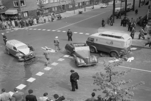 Eine Schwarz-Weiß-Szene eines Autounfalls an einer Straßenkreuzung, mit mehreren Fahrzeugen einschließlich eines Lieferwagens, einer Gruppe von Menschen drum herum und Gebäuden, Bäumen, Pfählen und Texttafeln im Hintergrund.