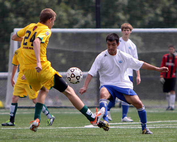 Spieler in gelben und weißen T-Shirts spielen Fussball auf einem Rasenplatz, mit einem Netz, Pfosten und Bäumen im Hintergrund.