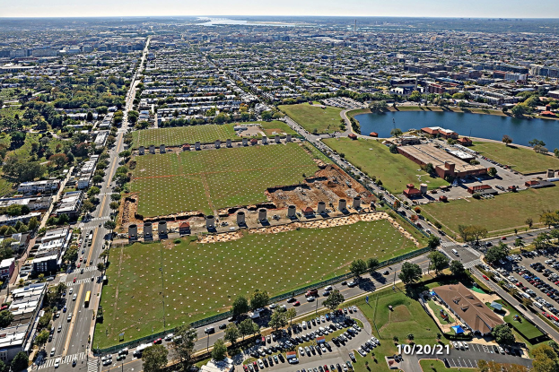 Luftaufnahme einer Stadt mit Gebäuden, Bäumen, Gras, Straßen mit Fahrzeugen, Wasser und bewölktem Himmel, die einen großen zentralen Platz als Parkstelle zeigt.