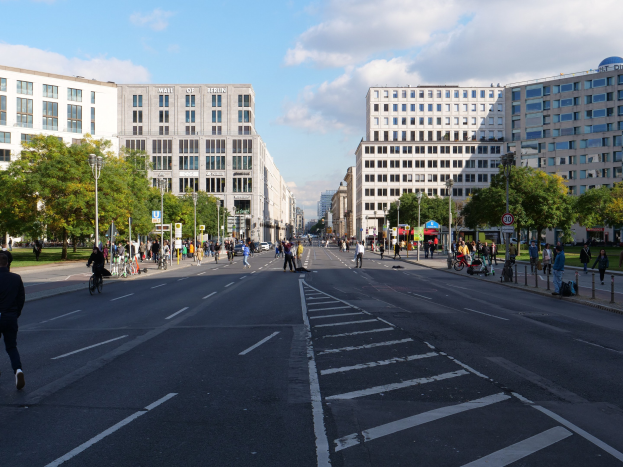Eine belebte Straßenkreuzung in Berlin, Deutschland, mit Fußgängern und Radfahrern auf der Straße, umgeben von hohen Gebäuden, Bäumen, Laternenmasten und Schildern unter einem bewölkten Himmel.