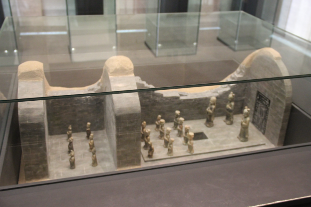 Ein Museumsglasvitrine mit verschiedenen Artefakten, darunter Skulpturen und Glaskästen, vor einer Wand.