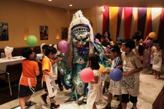 Eine Gruppe von Kindern steht um eine Person in einem Kostüm mit Ballons herum, mit einer Frau mit einer Kamera im Hintergrund, in einem Raum mit Stühlen, Tischen, Fotorahmen an der Wand und einer beleuchteten Decke.
