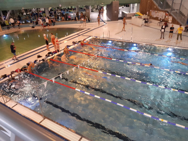Großes Hallenbad mit schwimmenden, stehenden, sitzenden und Gegenstände haltenden Menschen, mit Treppen, Geländern und einer Wand im Hintergrund.