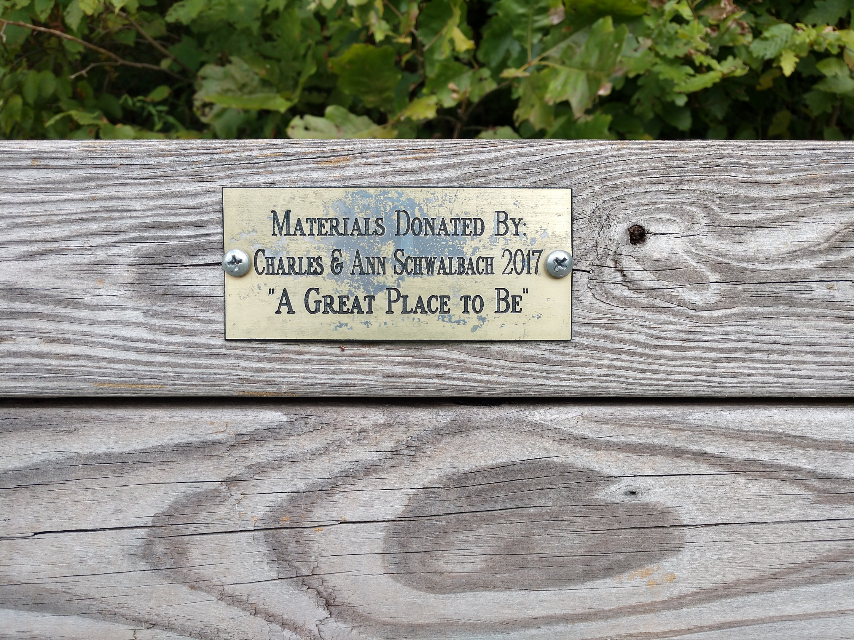 Holzbank mit einer Plakette, auf der "Materialien gespendet von Charles & Ann Schwabach 2017: Ein schöner Ort zu sein" steht, umgeben von üppigen grünen Pflanzen.