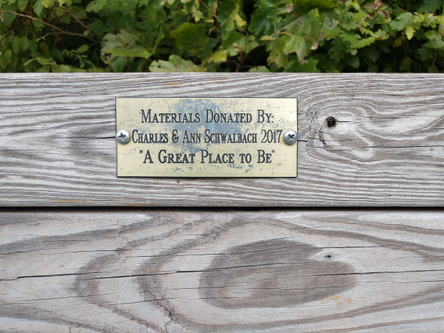 Holzbank mit einer Plakette, auf der "Materialien gespendet von Charles & Ann Schwabach 2017: Ein schöner Ort zu sein" steht, umgeben von üppigen grünen Pflanzen.