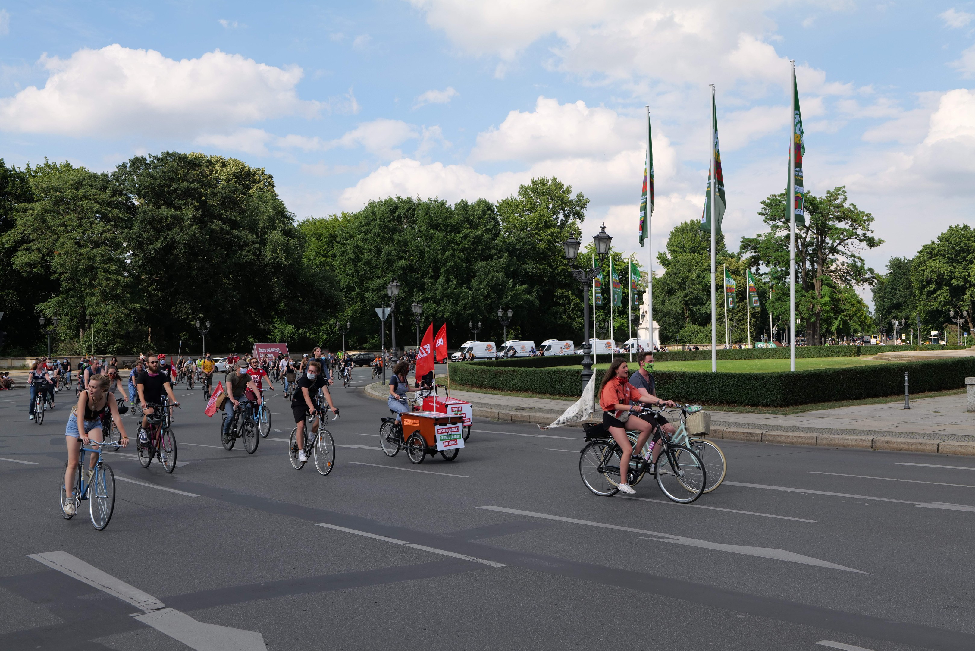 Eine Gruppe von Menschen, die auf Fahrrädern eine von Bäumen gesäumte Straße mit Laternenmasten und Flaggenmasten entlangfahren, während im Hintergrund Fahrzeuge und eine bewölkte Landschaft zu sehen sind.