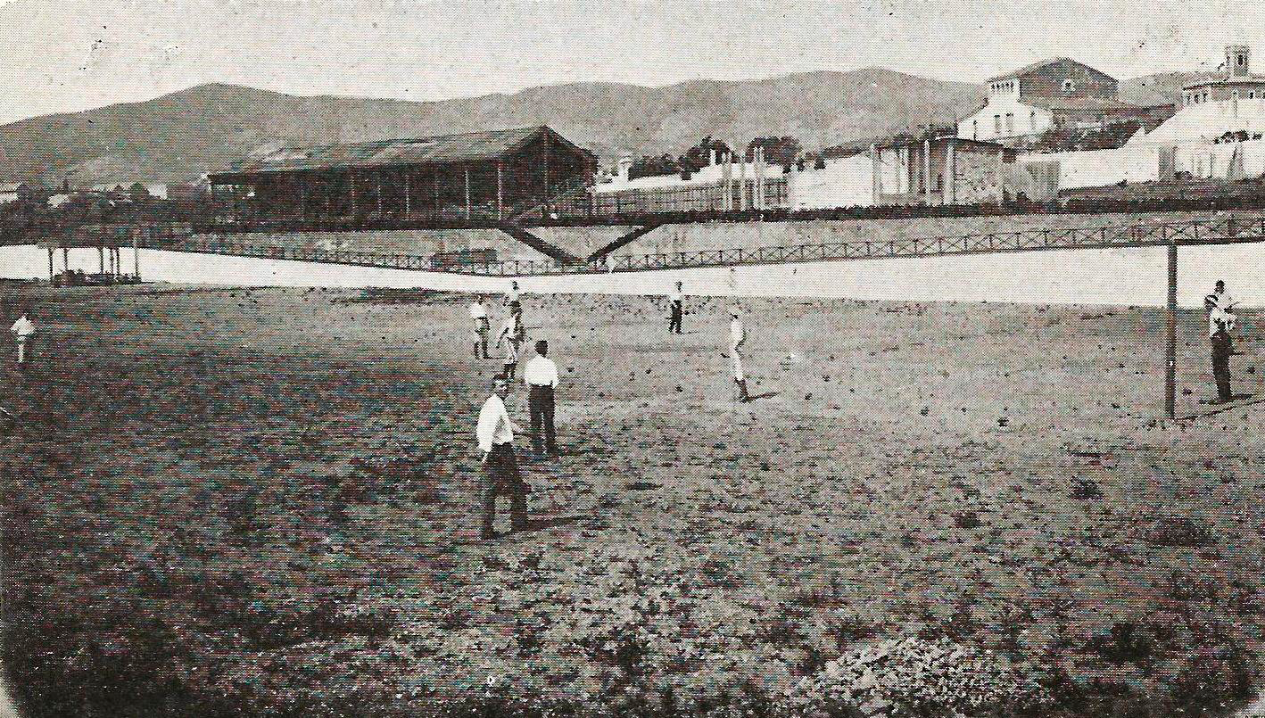 Ein Schwarz-Weiß-Foto einer Gruppe, die Cricket am Strand in den frühen 1900er Jahren spielt, mit Gebäuden, Bäumen, Hügeln und einer Brücke im Hintergrund und Text unten.