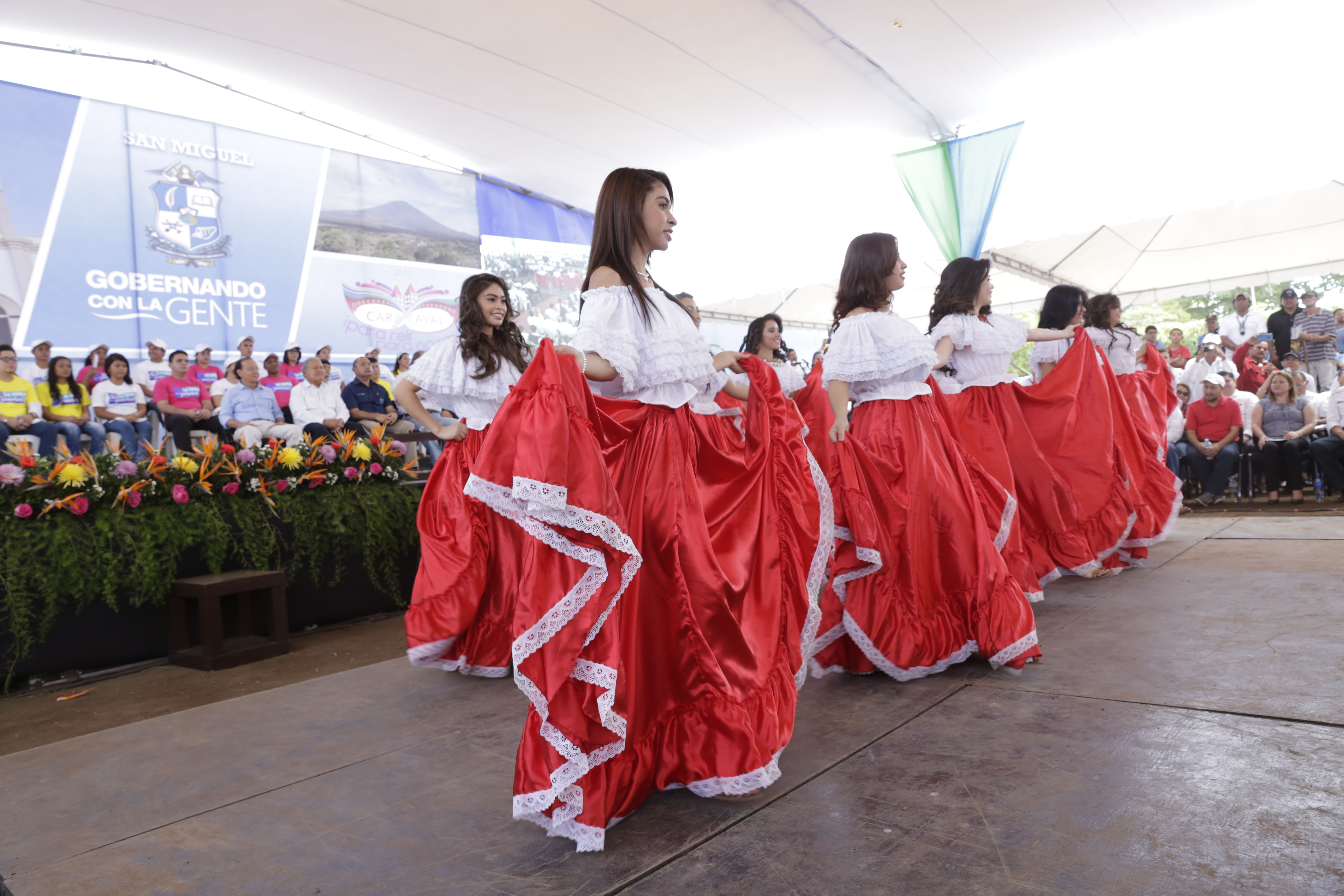 Eine Gruppe von Frauen in roten Kleidern tanzt auf einer Bühne vor einem sitzenden Publikum, mit Bannern, Pflanzen und einem Zelt im Hintergrund.