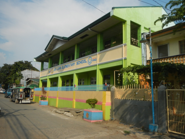 Ein Schulgebäude mit einer grünen und gelben Fassade, einem Zaun mit einem Tor, Topfpflanzen, Fahrzeugen auf der Straße, einem Straßenpfahl, Bäumen, Überlandleitungen und einem bewölkten Himmel.