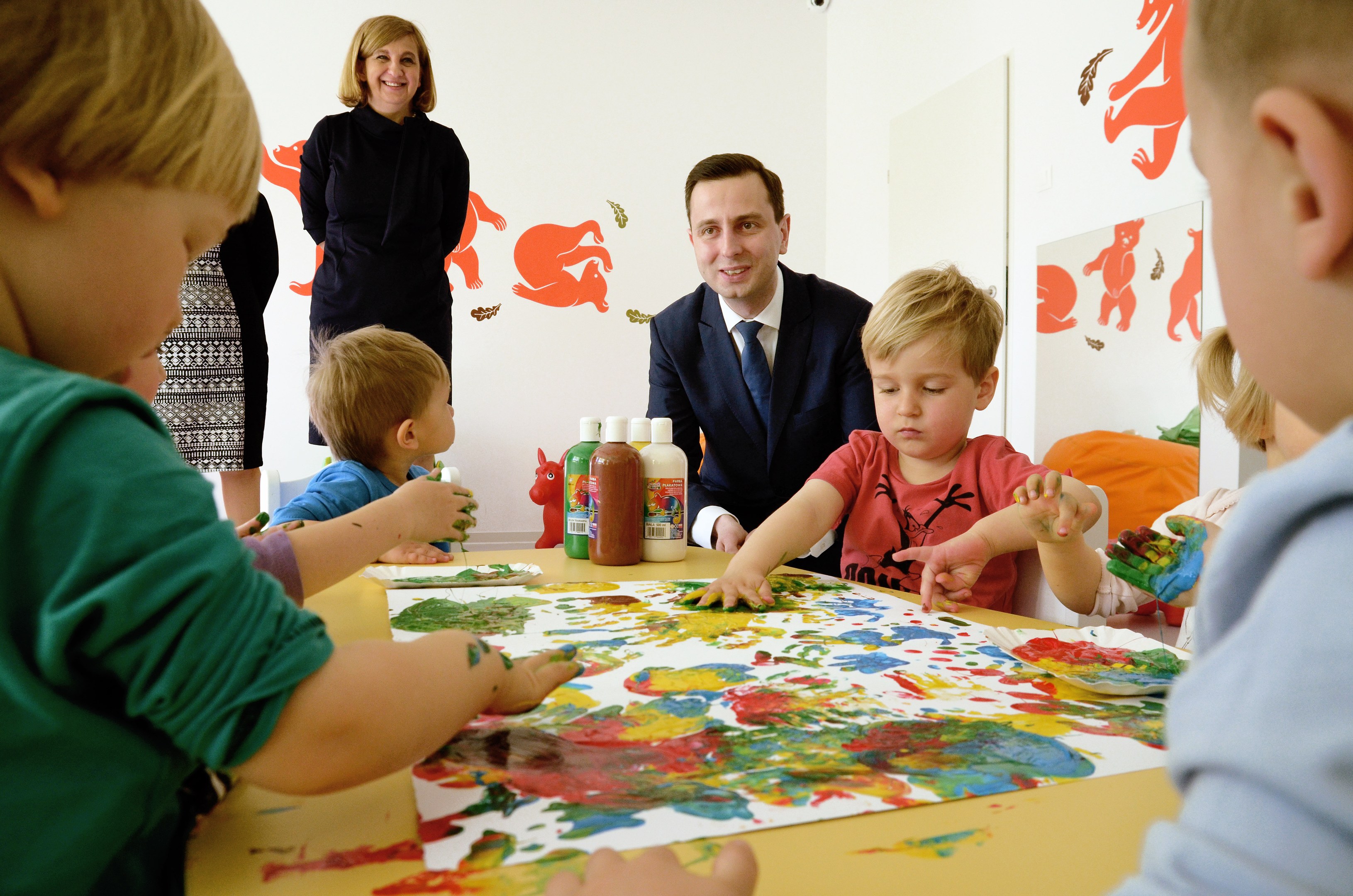 Kinder sitzen um einen Tisch mit Farbe an den Händen, beschäftigt mit einer kreativen Aktivität mit Papieren und Flaschen auf dem Tisch, während eine Frau im Hintergrund lächelt.