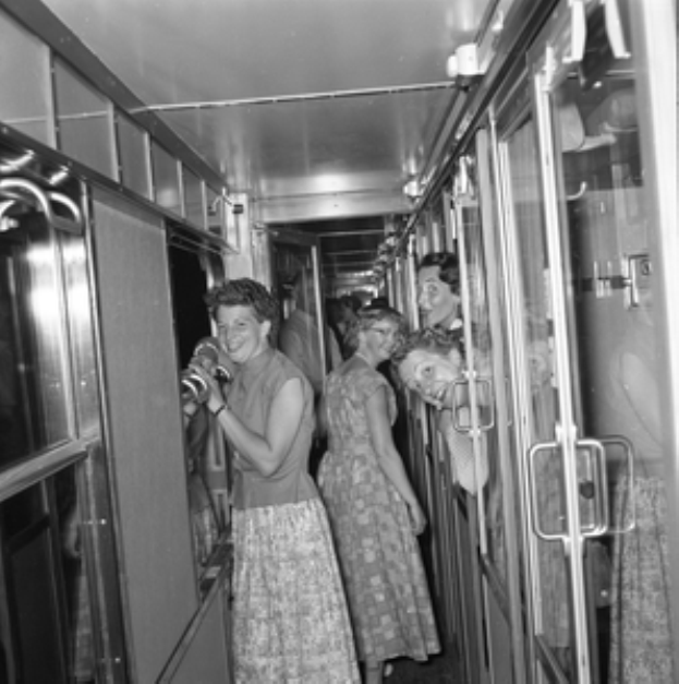 Eine Gruppe von Frauen, die auf einem Zug mit Türen auf beiden Seiten und Lichtern oben steht, dargestellt in Schwarz-Weiß.