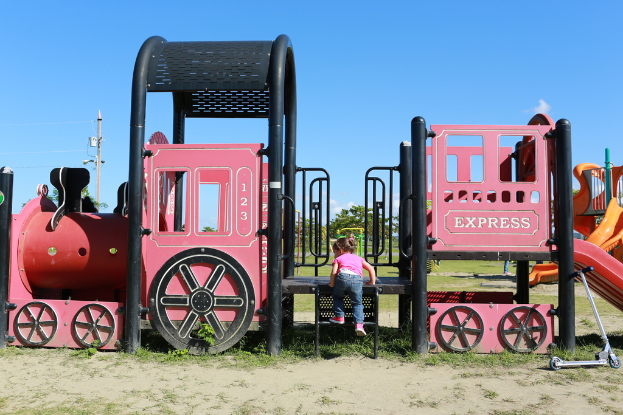 Ein Kind steigt auf einer Spielplatzanlage mit Bäumen, Polen, Stromleitungen und Wolken im Hintergrund und einem Roller auf dem Boden rechts davon.