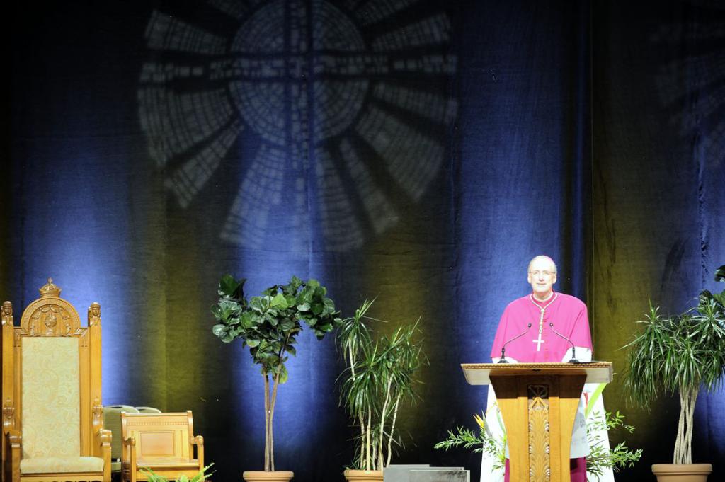 Ein Mann in einem rosafarbenen und weißen Kleid steht neben einem Podium in einem Auditorium, mit Pflanzen und zwei Stühlen daneben und einer bunten Wand im Hintergrund.