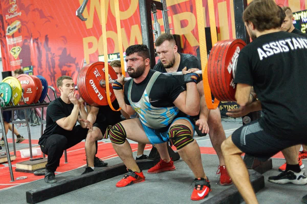 Eine Gruppe von Männern in einem Fitnessstudio, die Kniebeugen mit Hanteln machen, umgeben von verschiedenen Fitnessgeräten und Bannern mit der Aufschrift "Powerlifting-Weltmeisterschaften 2019".