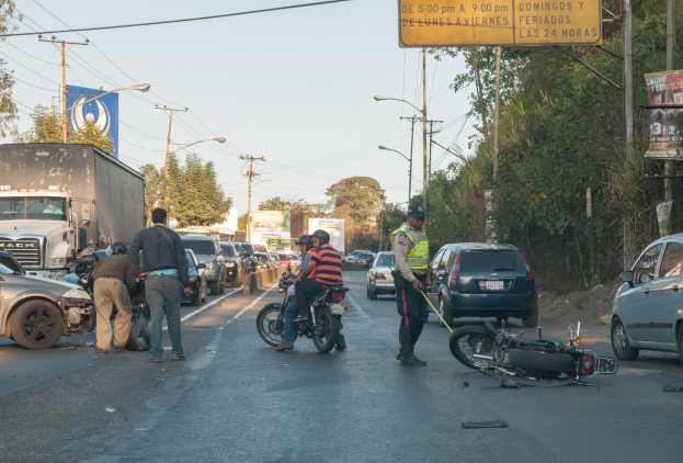 Eine Gruppe von Menschen steht um ein verunglücktes Motorrad auf der Straßenseite mit mehreren Fahrzeugen, darunter ein Lastwagen, und Hintergrundelementen wie Bäumen, Mästen, Lichtern und Schildern.