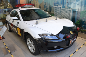 Polizeiauto vor einer Glaswand geparkt, mit einem Rad sichtbar, auf einem Boden neben einer Texttafel, mit Gebäuden, Bäumen und Himmel im Hintergrund sichtbar