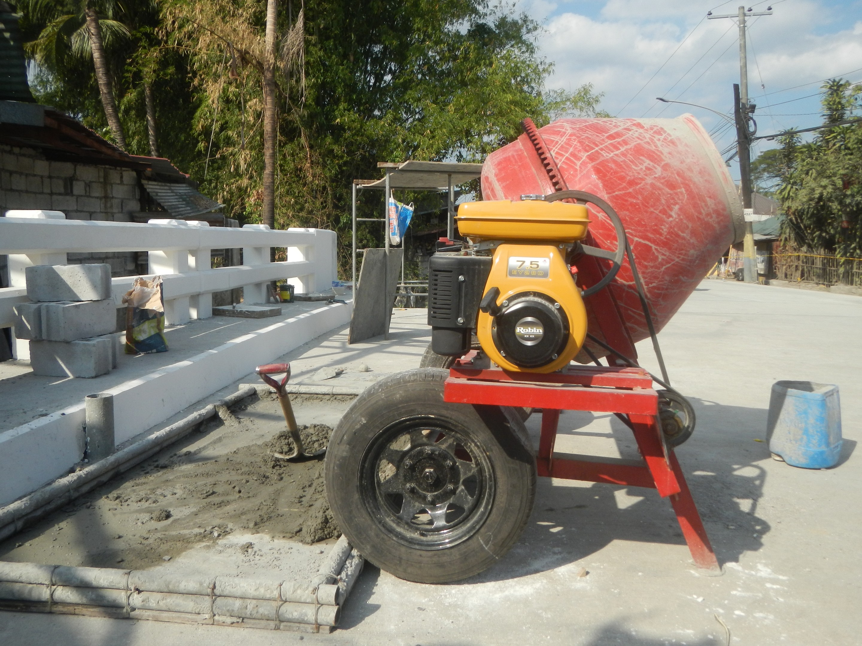 Betonmischer auf einem Zementlaster auf der Straße mit Betonblöcken, einem Eimer, einem Handwagen, einem Zaun, einem Haus, Bäumen, Strommasten mit Drähten und einem bewölkten Himmel.