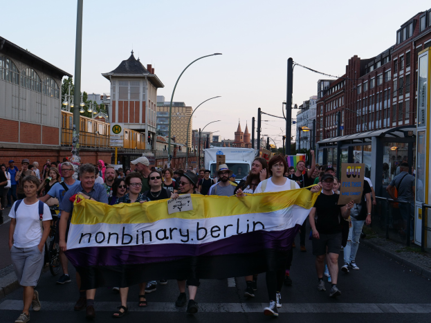 Gruppe von Menschen, die eine Straße in Berlin entlanggehen und ein "Nonbinary Berlin"-Schild halten, mit Laternenpfählen, Gebäuden, einem Bushaltestellenschild, Bäumen und einem klaren blauen Himmel im Hintergrund.