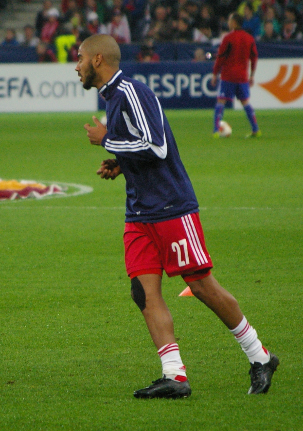 Ein Mann in einem blauen Hemd und roten Shorts läuft auf einem Fußballfeld, im Hintergrund sitzen einige Menschen und es sind einige Werbetafeln zu sehen.
