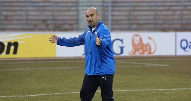 Ein Mann in einer blauen Jacke steht auf einem Fußballfeld, umgeben von Schildern mit Text und Logos, entschlossen.