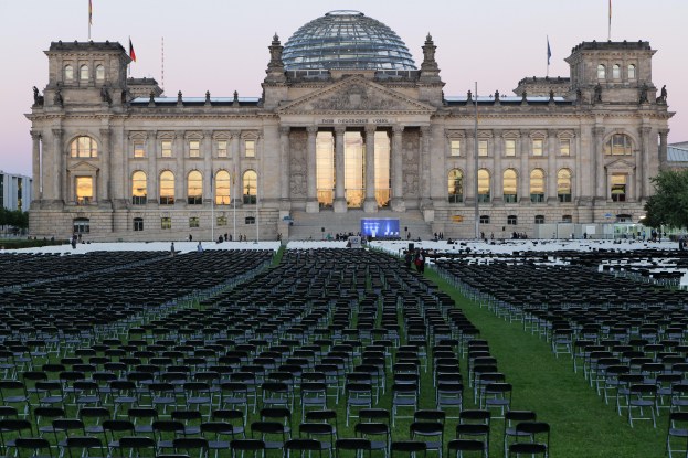 Das Reichstagsgebäude in Berlin, Deutschland, mit zahlreichen Stühlen davor, Fenstern, Säulen, Bögen und Fahnen, sowie einigen Menschen im Vordergrund und Bäumen unter einem klaren blauen Himmel im Hintergrund.