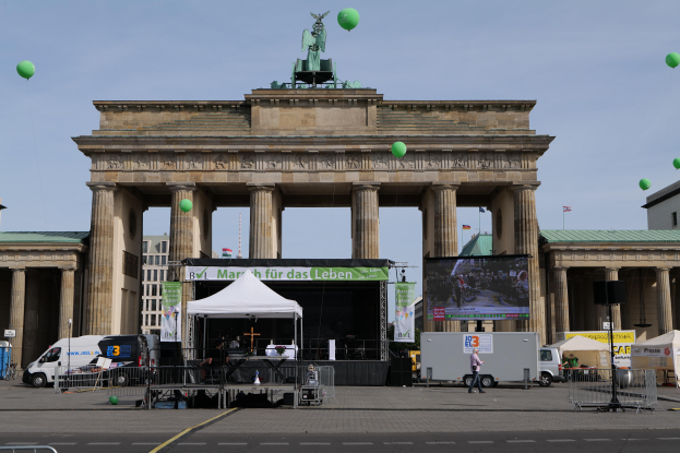 Das Brandenburgertor in Berlin, Deutschland, mit einer Bühne davor, einigen Menschen auf der Bühne, parkenden Fahrzeugen in der Nähe, einem großen Bildschirm rechts, Gebäuden im Hintergrund und grünen Ballons am Himmel.