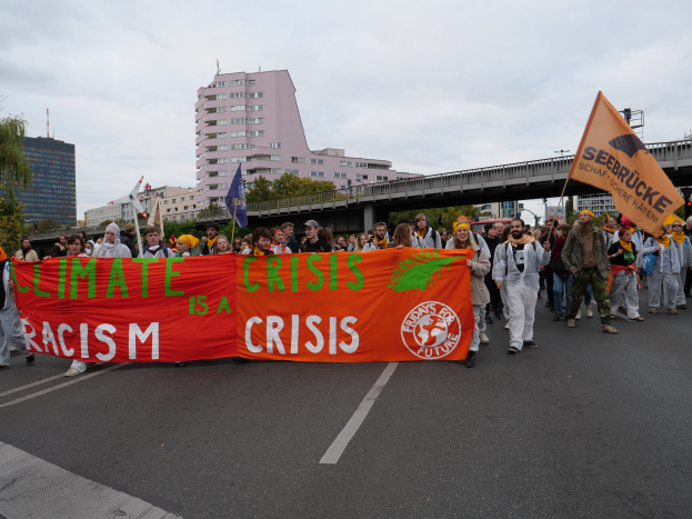 Eine Gruppe von Menschen marschiert eine baumbestandene Straße entlang und hält ein Banner hoch, auf dem "Klima-Krise ist eine Krise" steht, mit Gebäuden und einer Brücke im Hintergrund unter einem bewölkten Himmel.