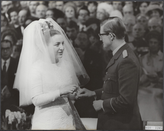 Ein Schwarz-Weiß-Foto von einem Mann und einer Frau, die sich die Eheringe austauschen, mit einer Gruppe von Menschen im Hintergrund.