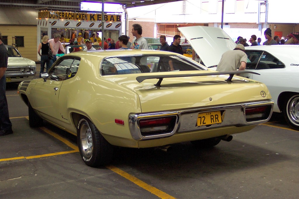 Ein Oldtimer steht auf einer Straße mit Menschen in der Nähe, einem Geschäft in der Mitte und Gebäuden im Hintergrund.
