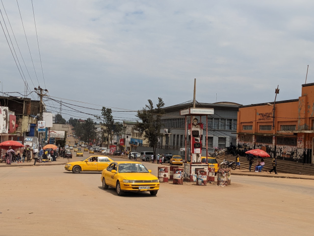 Ein gelbes Taxi fährt eine belebte Stadtstraße entlang, gesäumt von Gebäuden, wo Fußgänger mit Regenschirmen unter einem bewölkten Himmel auf dem Gehweg gehen, mit Strommasten und Drähten im Hintergrund.