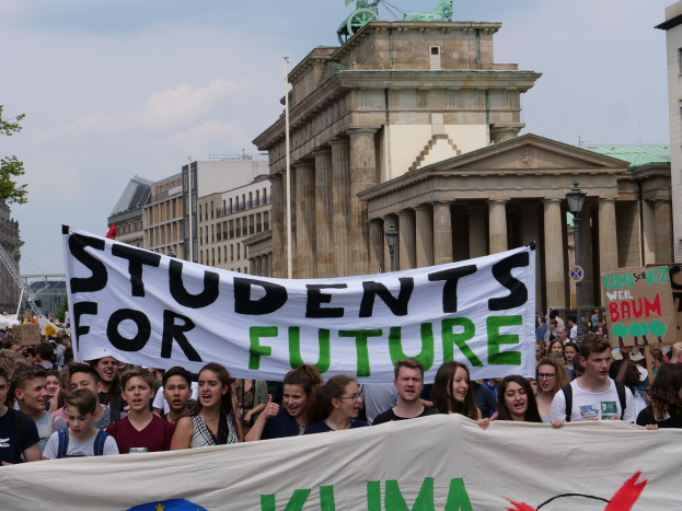 Gruppe von Sch├╝lerinnen und Sch├╝lern marschiert in Berlin mit einem bunten "Students for Future"-Schild vorbei an Geb├Ąuden, B├Ąumen und Himmel.