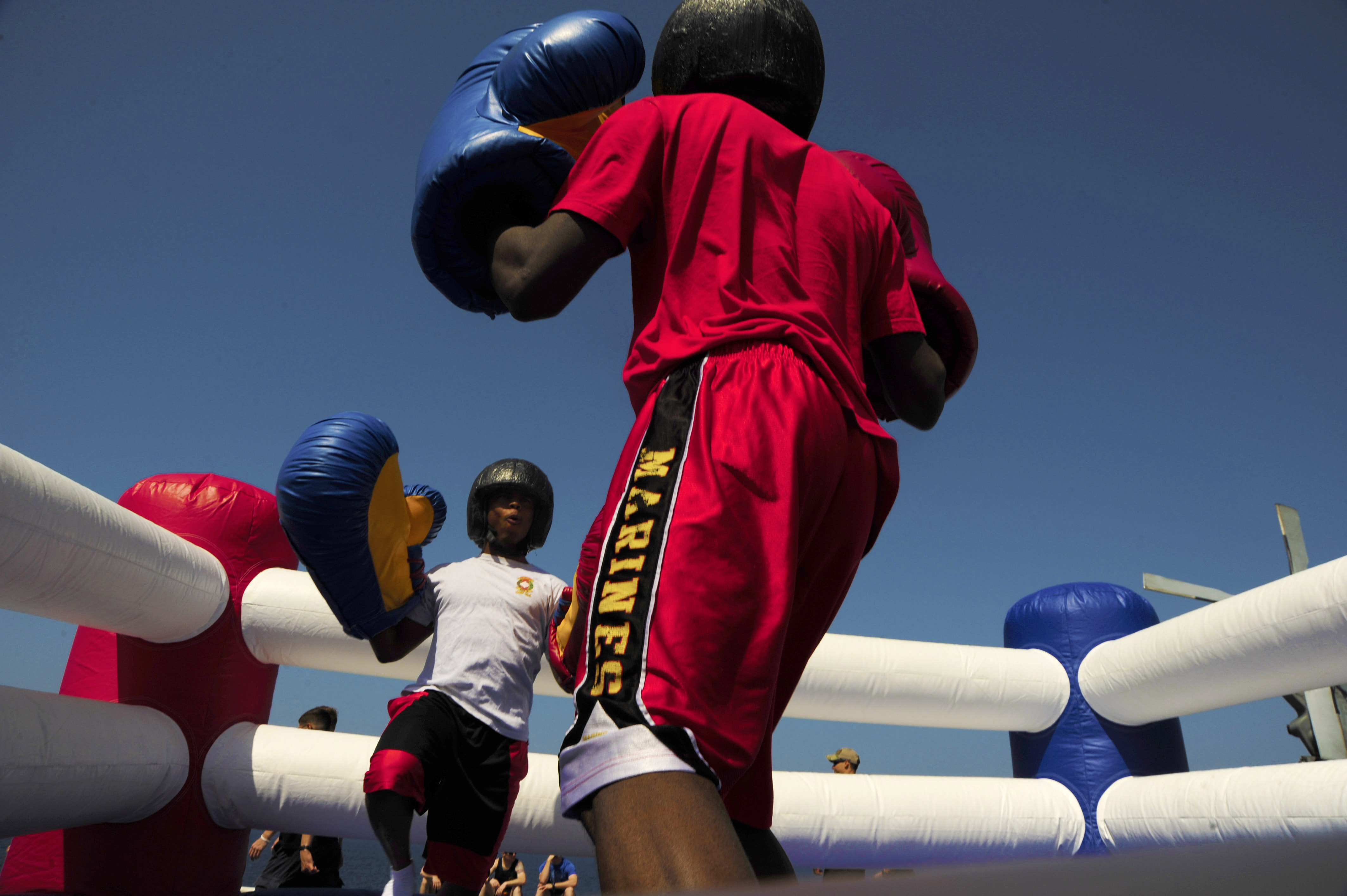 Zwei Boxer in einem Boxring, einer in rotem Hemd und blauen Shorts, kämpfen mit Boxhandschuhen, umgeben von ein paar Menschen und unter einem klaren blauen Himmel.
