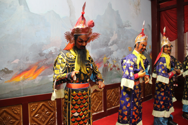 Eine Gruppe von Menschen in Kostümen steht zusammen auf einem roten Teppich, mit einem Gemälde an der Wand dahinter und einem Vorhang auf der rechten Seite.
