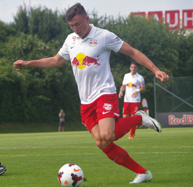 Ein professioneller Fußballspieler in einem roten Trikot tritt einen Ball auf einem grünen Feld mit Bäumen und einem klaren blauen Himmel im Hintergrund, mit "RB Leipzig" auf einem Schild.