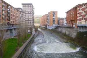 Ein Fluss fließt durch eine Stadt mit hohen Gebäuden auf beiden Seiten, umgeben von grünem Gras, Bäumen und Laternenmasten, überquert von einer Brücke, mit Hügeln und einem klaren blauen Himmel im Hintergrund.