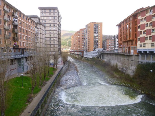 Ein Fluss fließt durch eine Stadt mit hohen Gebäuden auf beiden Seiten, umgeben von grünem Gras, Bäumen und Laternenmasten, überquert von einer Brücke, mit Hügeln und einem klaren blauen Himmel im Hintergrund.