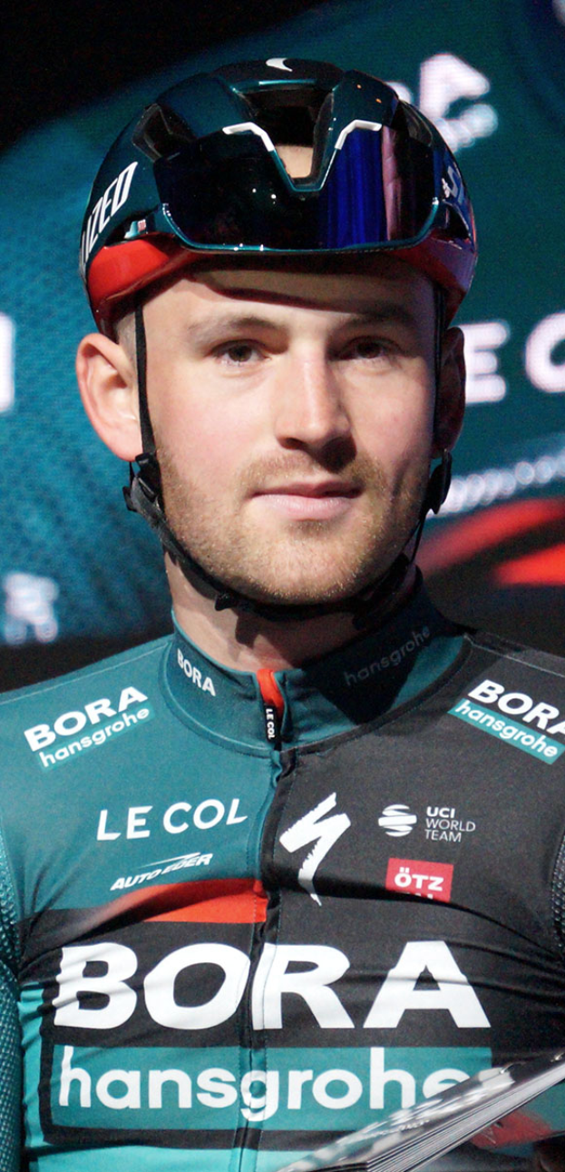 Ein Mann in einem blauen Helm und einem Trikot steht neben einem Fahrrad, mit einem 'Bora-Hansgrohe World Tour'-Plakat im Hintergrund und entschlossener Miene.