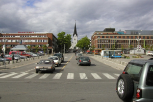 Stadtstraße mit parkenden Autos, Gebäuden, Bäumen, Laternen, Wolken am Himmel und einem sichtbaren Kfz-Kennzeichen.