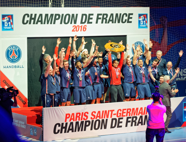 Eine Gruppe von Männern auf einer Bühne mit einem Pokal, einer Person filmt und einem "Champion de France 2016"-Schild im Hintergrund.